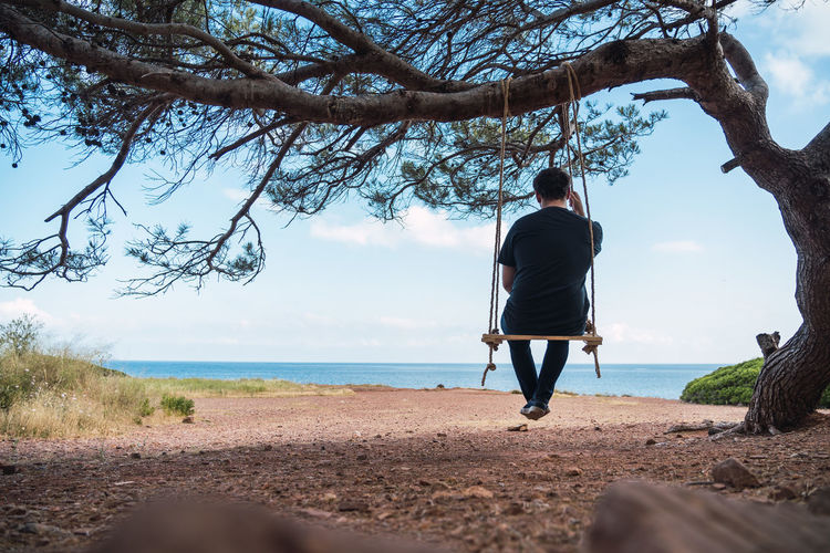 Rear view of man sitting on rope swing against sea