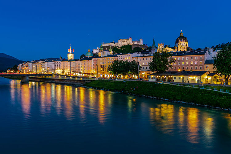 Illuminated buildings by river against blue sky