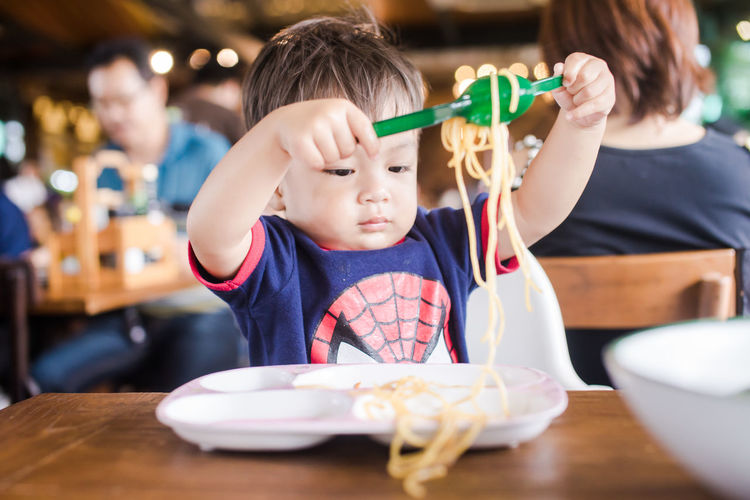 Cute baby boy playing with noodles at table in restaurant