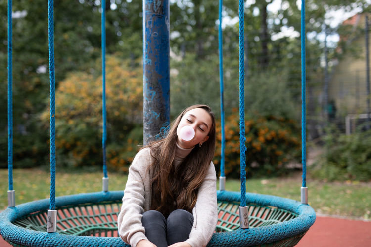 Smiling young woman blowing bubblegum while sitting in swing at playground
