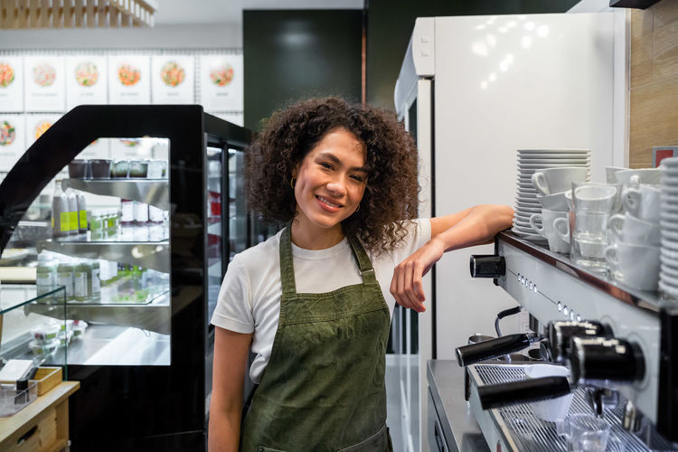 Delighted ethnic female barista in apron and with curly hair leaning on coffeemaker while standing in cafe and looking at camera