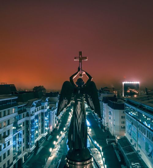 Angel statue with cross against sky in city at night