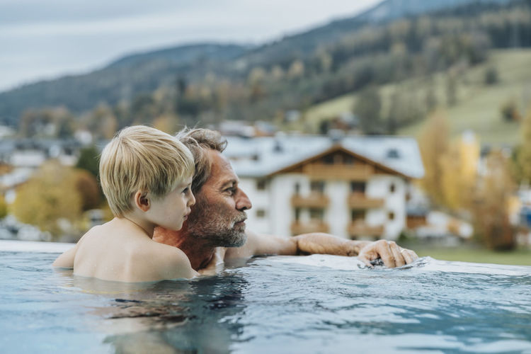 Father and son in infinity pool looking away at salzburger land, austria