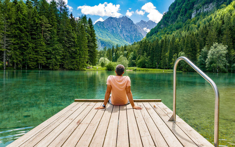 Rear view of young man sitting on deck by lake in mountains.