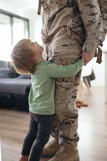 Side view of cute son greeting and embracing legs of crop anonymous military parent in camouflage uniform after coming home from mission