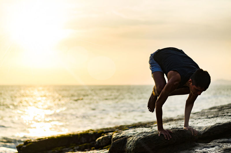 Full length of man doing yoga on rock at beach during sunrise