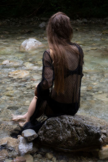 Rear view of woman sitting on rock by river