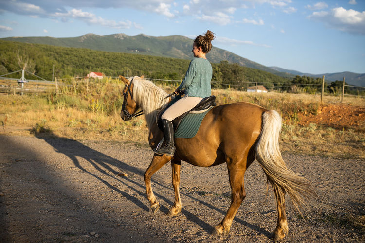 Back view of female riding stallion with smooth brown coat on rough land against mount in countryside