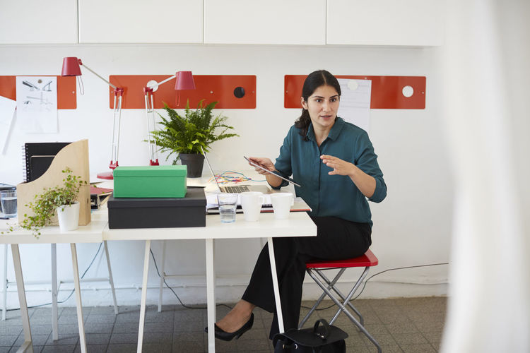 Mid adult businesswoman gesturing while holding digital tablet at table in office