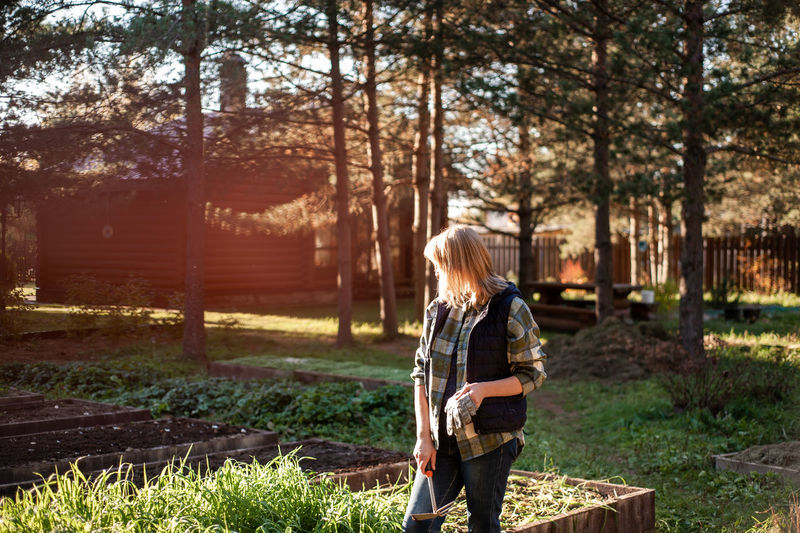 A blonde woman  with gloves and a rake in her hands stands in the backyard in the sunset 