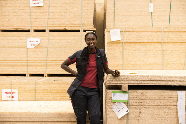 Portrait of smiling saleswoman with hand on hip standing near plywood stack at hardware store