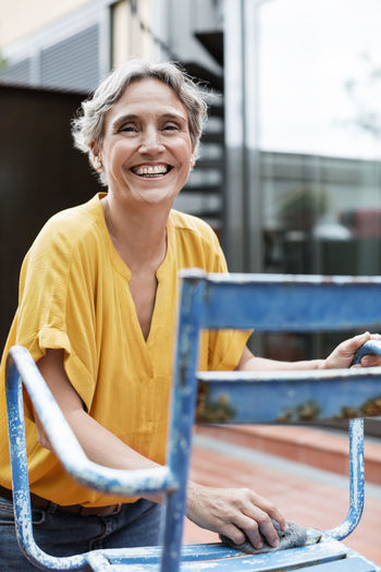 Portrait of cheerful mature woman scrubbing metallic chair with steel wool at yard