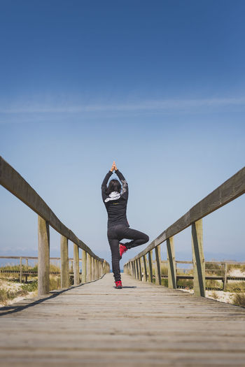 Rear view full length of woman doing yoga on pier against blue sky