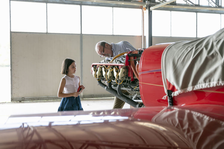 Grandfather concentrating while showing how to fix airplane tool to granddaughter while standing in hangar