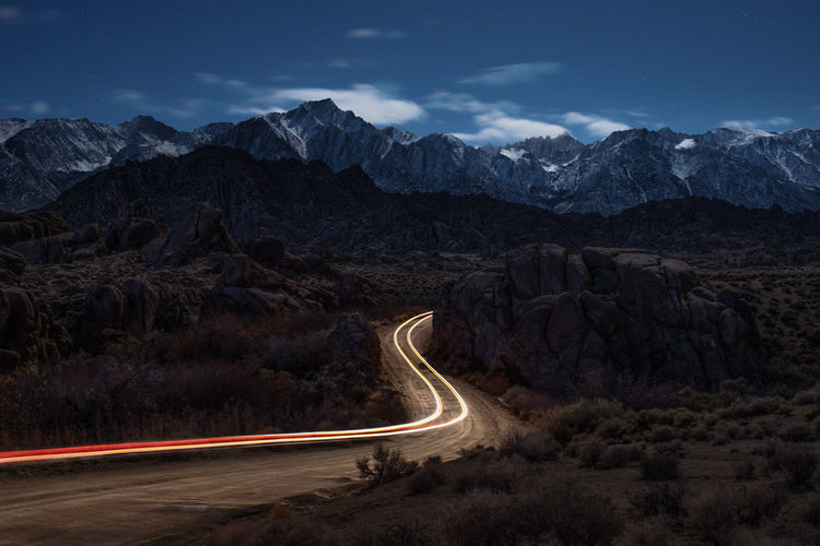 Light trails on road by mountains against sky at night