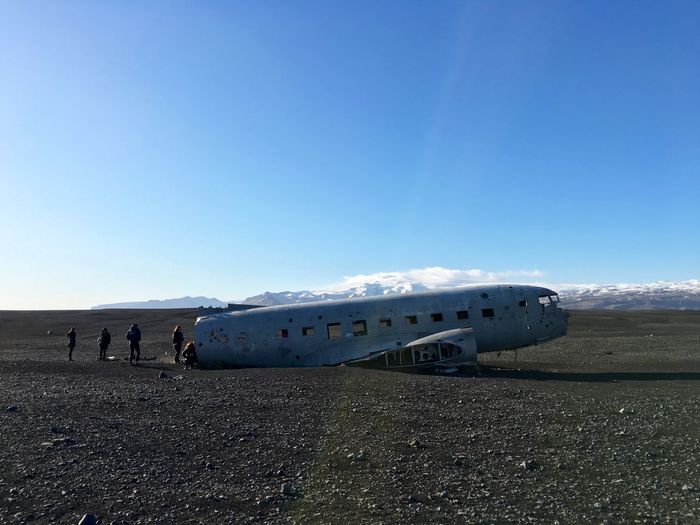 People by crashed airplane wreck on black sand against blue sky