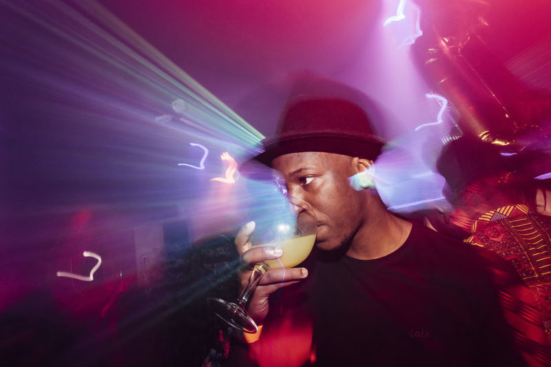 Low angle of african american guy sipping alcohol cocktail under colorful illumination during party in nightclub