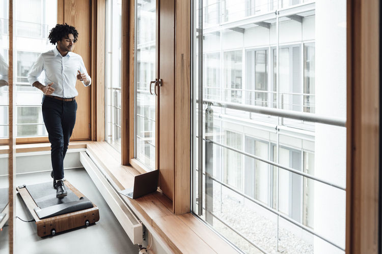 Active male entrepreneur walking on treadmill while looking at laptop against window in workplace