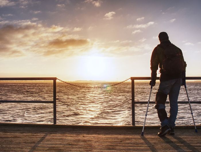 Hurt man hiker walk with forearm crutch on wharf mole above sea to sun. fantastic silent morning