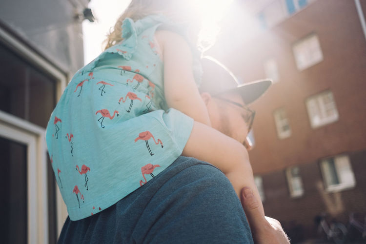 Low angle view of father carrying daughter on shoulders in city during sunny day