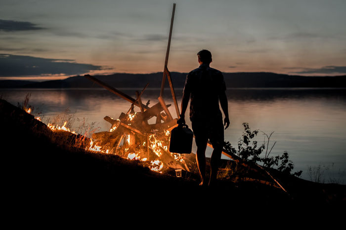 Rear view of man standing in front of campfire by lake