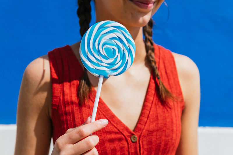Crop of anonymous female standing with sweet lollipop in street on sunny day on blue background and looking away