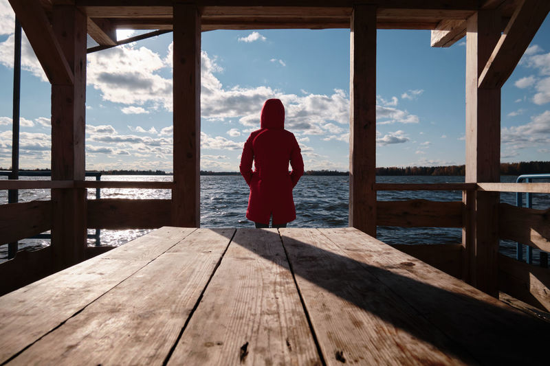 A woman in a red jacket sits at a table on a wooden pier and looks at the water