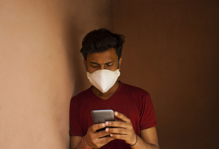 Close-up of young man wearing flu mask using smart phone against wall at home