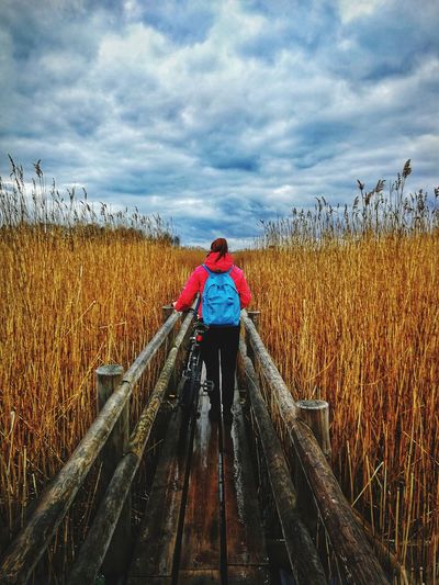 Rear view of female hiker walking with bicycle on footbridge amidst field against cloudy sky