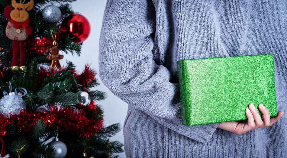 Midsection of woman holding christmas gift in hand behind back standing by christmas tree at home