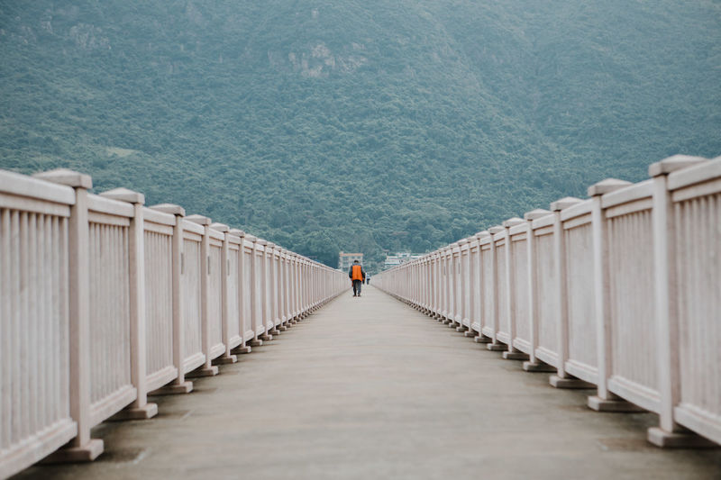 Man wearing orange jacket walking in center of bridge with a vanishing point