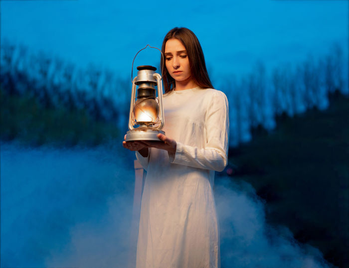Young woman with illuminated oil lamp standing in forest at dusk