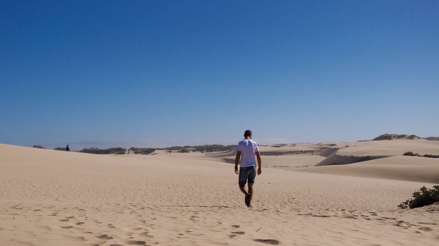 Rear view of man walking on sand in desert against clear blue sky