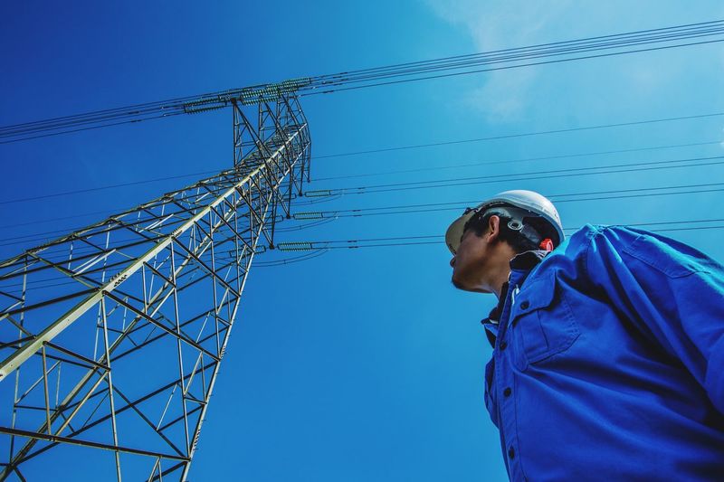 Low angle view of man standing by electricity pylon against clear blue sky