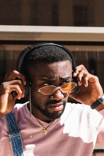 Cool african american male hipster in trendy outfit standing on street and listening to music in wireless headphones