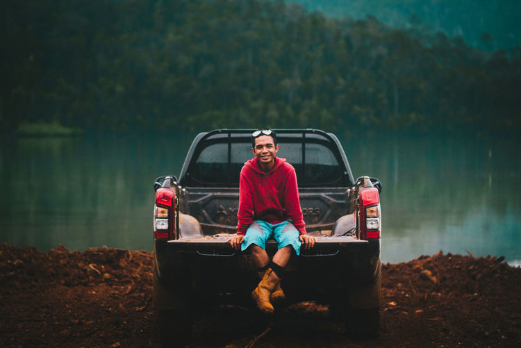 Full length portrait of young man sitting by lake