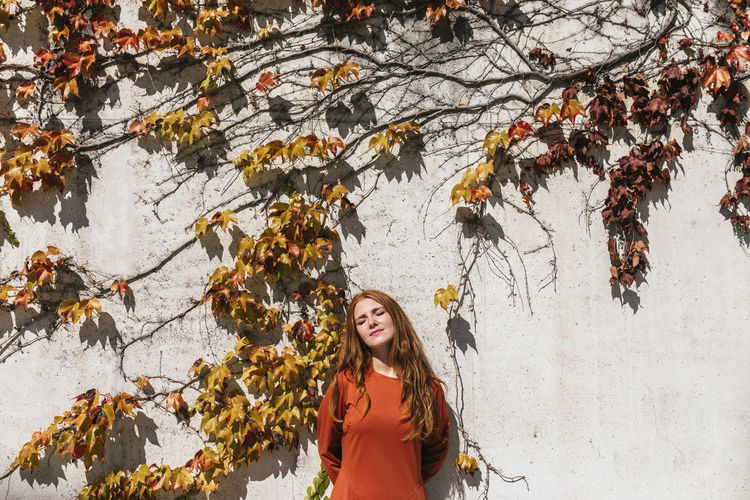 Woman with eyes closed and hands behind back standing against ivy wall