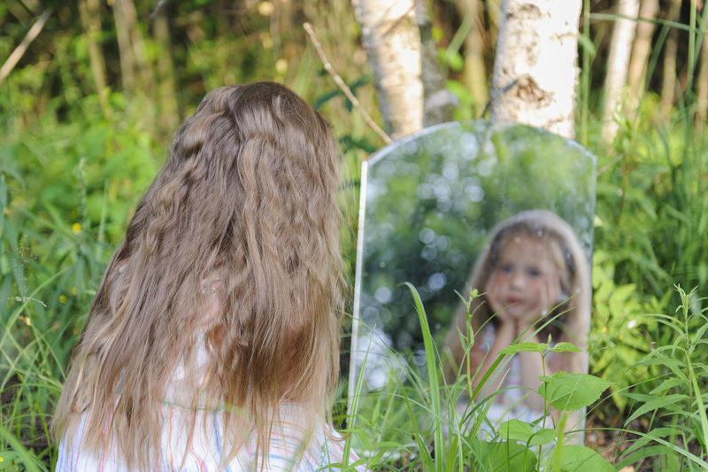 Rear view of girl with long hair looking in mirror at forest