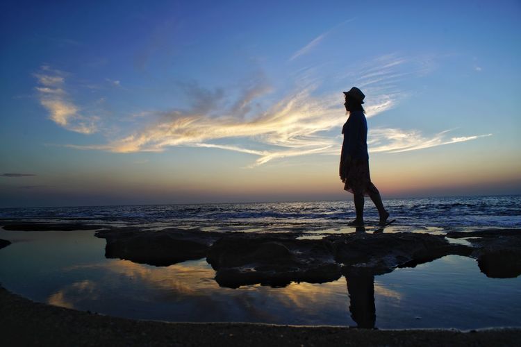 Side view of silhouette woman standing at beach against sky during sunset