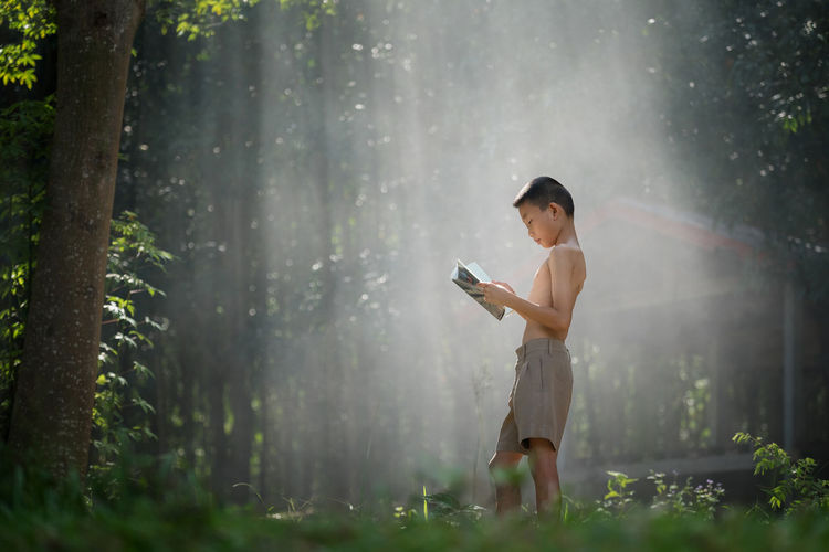 Side view of shirtless boy reading book while standing on field