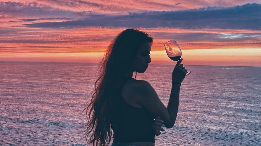 Woman drinking water at beach against sky during sunset