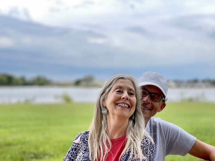 Happy senior couple sitting against river, grass and stormy sky.  portrait with red and blue tops.