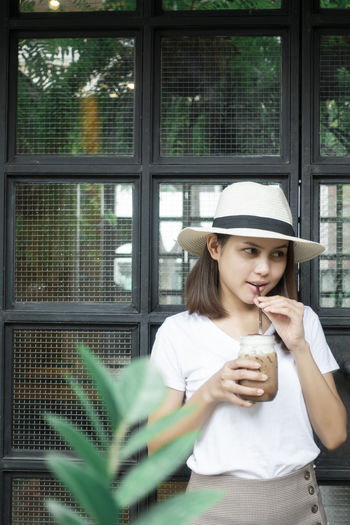Smiling young woman drinking iced coffee against window