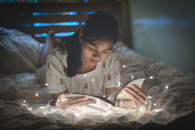 Young woman is reading a book with led lights on the bed in her bedroom.