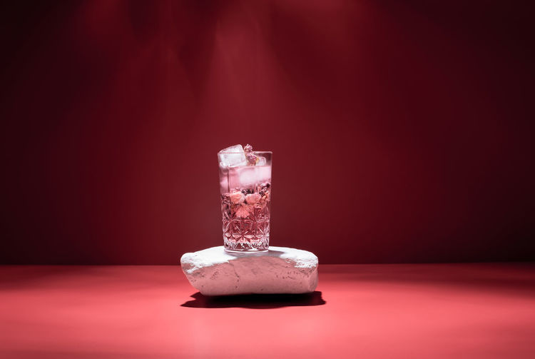 Glass of pink alcohol cocktail with ice cubes on white stone against red backdrop in studio