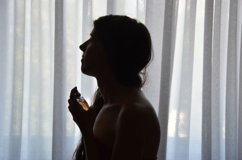 Side view of woman spraying perfume against curtain