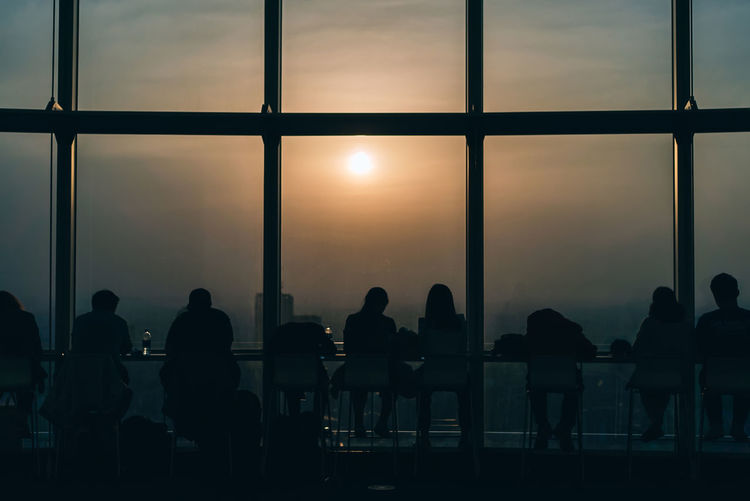 Silhouette people sitting by glass window at sunset