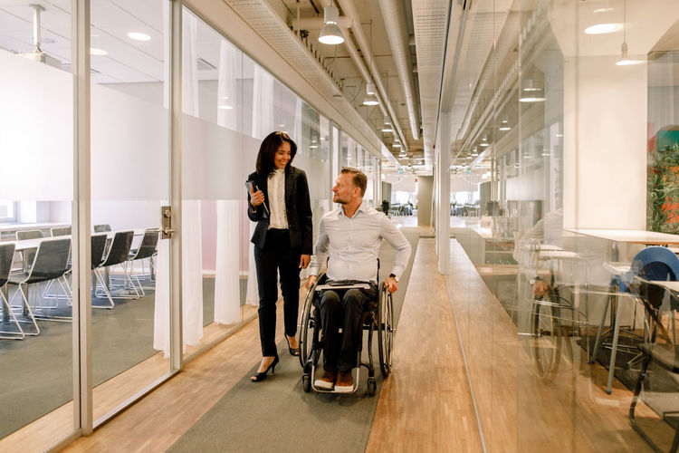 Smiling businesswoman looking at disabled colleague sitting on wheelchair in corridor at work place