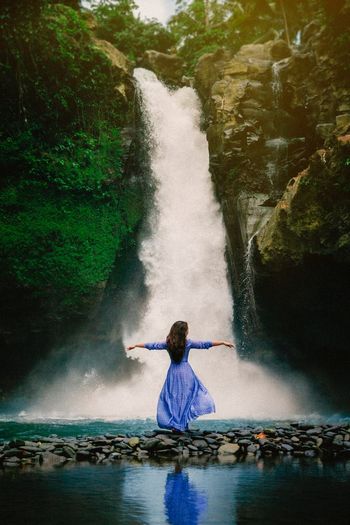 Rear view of woman with arms outstretched standing against waterfall in forest