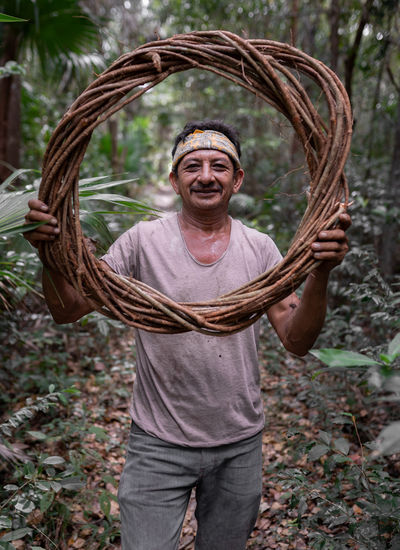 Positive male worker looking at camera through round woven tree branches while standing in nature with trees on summer day